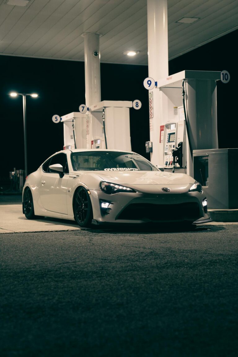A sleek white sports car parked at a gas station pump during the night, under bright lighting.